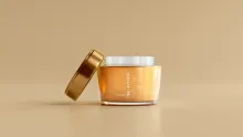 Tarro de crema de vidrio con tapa dorada apoyada, sobre fondo beige liso